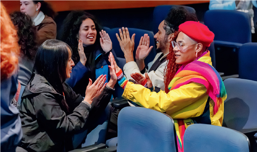 
Descrição da imagem: Fotografia horizontal de Bárbara, Luna, Amanza e Pedro sentados em cadeiras azuis do auditório.
Bárbara está à esquerda na primeira fileira, de frente para Luna, que está na segunda fileira à direita. As duas estão com as mãos erguidas e espalmadas, uma em direção à outra; a palma da mão esquerda de Bárbara está encostada na palma da mão esquerda de Luna. Bárbara é negra de pele clara, com cabelos longos pretos e lisos, franja, sobrancelhas finas e nariz com a ponta arredondada. Usa um casaco preto.
Luna está com box braids ruivas, lateral esquerda raspada, boina vermelha, casaco amarelo e um lenço comprido com as cores da bandeira LGBTQIAPN+ sobre o ombro esquerdo.
Também na segunda fileira, na cadeira do lado de Luna, Amanza está à esquerda e Pedro à direita. Estão com as mãos erguidas e espalmadas, uma em direção a outra. Os cabelos longos e pretos de Amanza estão parcialmente presos e ela usa casaco azul. Pedro está com os cabelos pretos presos em um coque no alto da cabeça e usa casaco branco. Fim da descrição da imagem.
