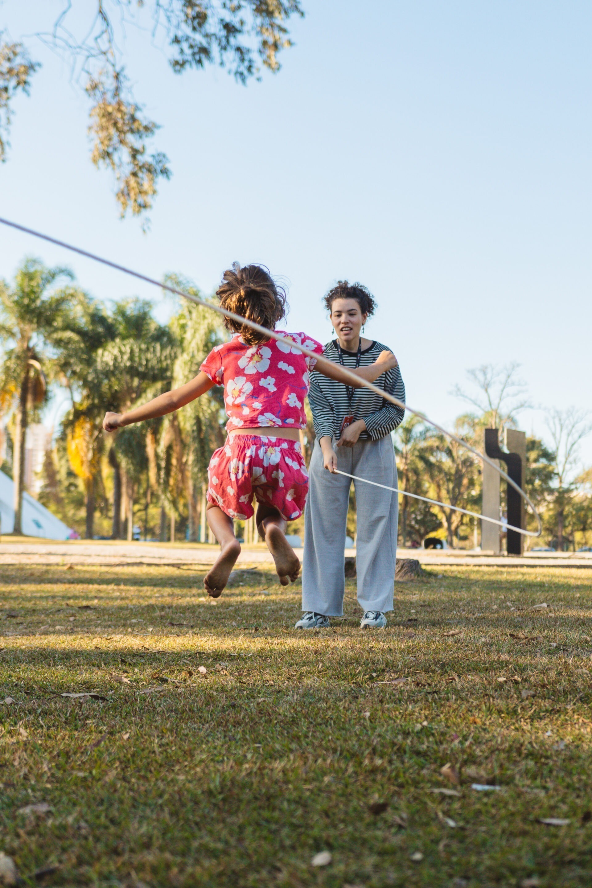 Descrição da imagem: Fotografia diurna horizontal de uma menina e da educadora Amanda Santos no Jardim de Esculturas em um gramado esverdeado. Sob o céu azul, ao centro, a menina é vista de costas. Ela é parda, de cabelos castanhos lisos presos em um rabo-de-cavalo. Usa um conjunto de camiseta e shorts de pano cor de rosa com estampa de flores brancas com o miolo amarelo e caule azul-escuro. A menina está pulando corda, parada no ar, com as pernas flexionadas e os braços abertos para as laterais.
Diante da menina, Amanda Santos segura a corda, que está parada no ar, projetada para a lateral direita. Amanda é uma mulher negra de pele clara, tem cabelos pretos cacheados e amarrados em um coque no meio da cabeça, sobrancelhas finas, nariz arredondado na ponta e lábios médios. Usa camiseta de mangas longas com listras horizontais em branco e preto, calça de moletom cinza e tênis brancos com detalhes em azul. Ao fundo, há árvores altas de copas esverdeadas. Fim da descrição da imagem.
