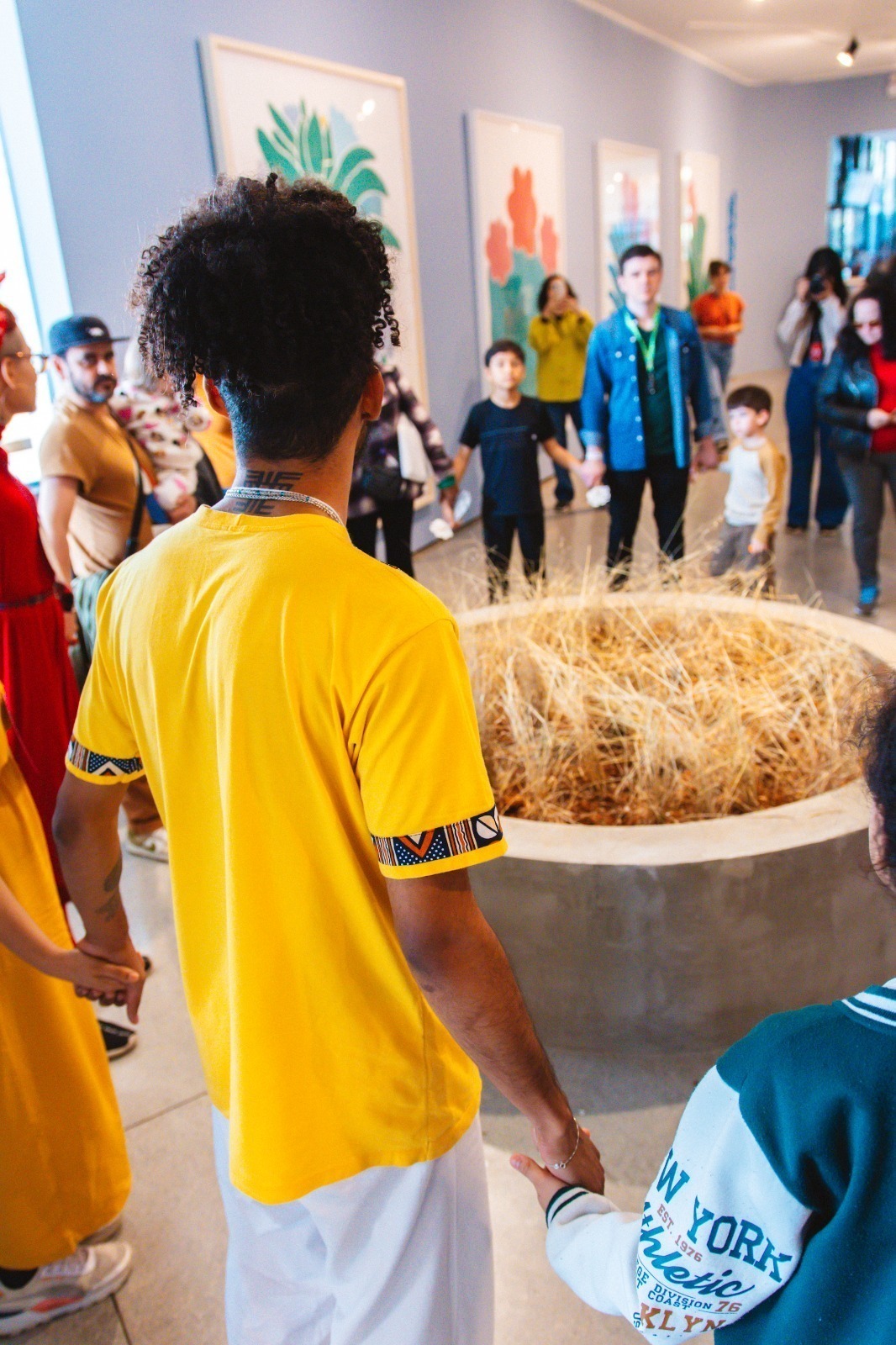 Descrição da imagem: Fotografia diurna vertical do educador Pedro com um grupo de pessoas, algumas enquadradas da cintura para cima e outras vistas de corpo inteiro, todas de mãos dadas e posicionadas ao redor da instalação composta pela mureta mediana de concreto, em formato circular e preenchida com os galhos secos.
À esquerda, Pedro é visto de costas. Ele é um homem negro de pele clara, magro, com os cabelos longos pretos cacheados amarrados em um coque alto e com a nuca raspada. Usa camiseta amarela e colar prateado. No grupo, há pessoas brancas e negras entre adultos e crianças.
O fundo está um pouco desfocado e, à esquerda, pendurados na parede azul-clara, quadros com pinturas de flores em tons de verde e Vermelho. Fim da descrição da imagem.
