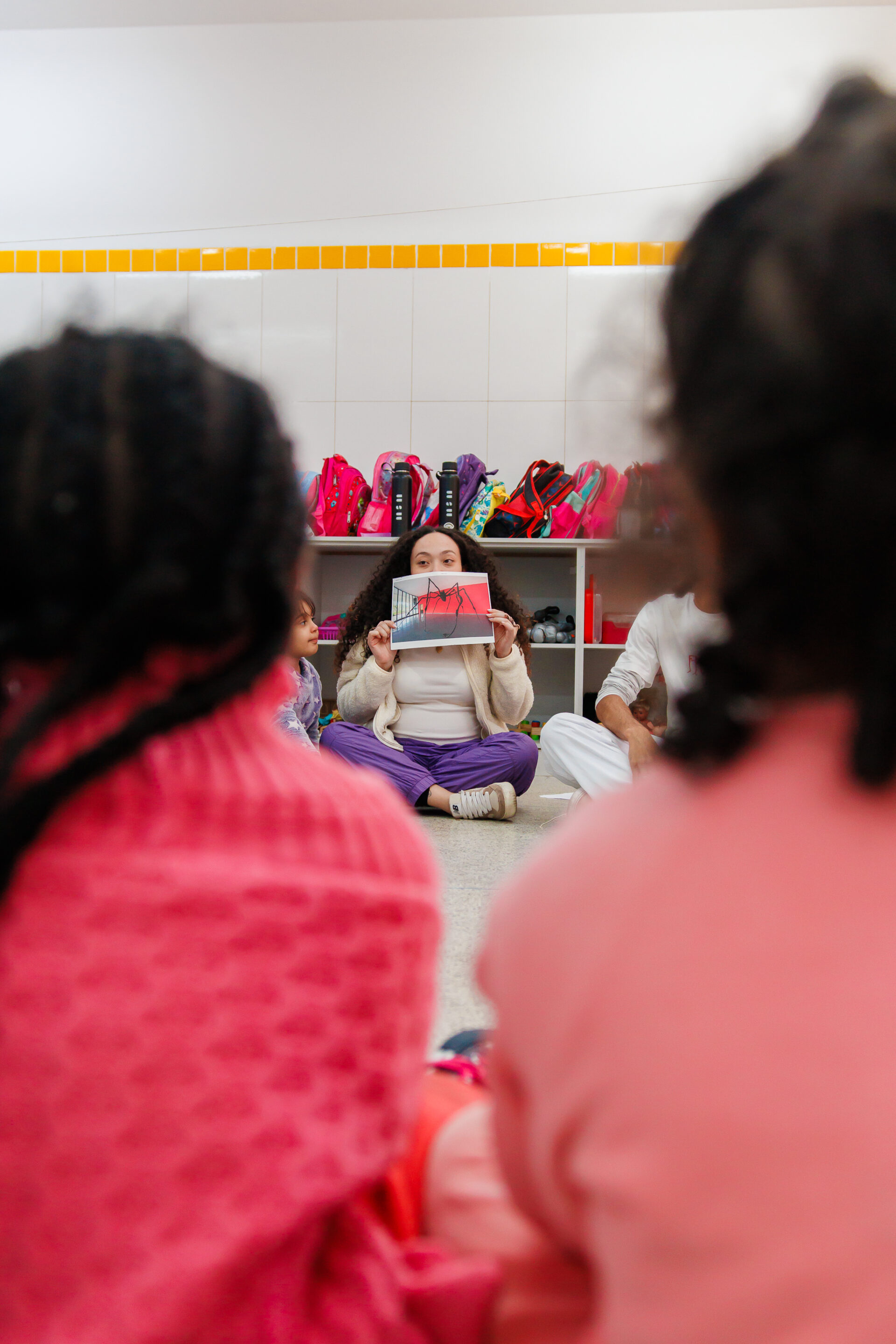 Descrição da imagem: Fotografia vertical da educadora Amanda Falcão com um grupo de crianças em uma sala com paredes e chão claros. Duas crianças são vistas parcialmente à frente, desfocadas, sentadas no chão de costas. Ambas têm cabelos pretos e usam roupas cor-de-rosa. Entre elas, em nitidez, Amanda Falcão é vista ao fundo e a esquerda, há outra criança parda com cabelos lisos. Amanda é uma mulher negra de pele clara, com cabelos longos, cacheados e volumosos repartidos ao meio, e sobrancelhas finas.
Amanda está sentada no chão, com as pernas cruzadas, segurando à frente do nariz e da boca uma fotografia de uma instalação do MAM: uma escultura de aranha de grandes dimensões, feita em material escuro. A aranha tem corpo segmentado e oito patas longas e finas, irregulares e inclinadas em diferentes direções. A peça está exposta em uma sala com a parede esquerda de vidro, a parede ao fundo vermelha e o chão claro.
Atrás de Amanda, há uma estante larga e baixa com prateleiras onde estão mochilas pequenas em tons de rosa, roxo, azul e verde. Fim da descrição da imagem.
