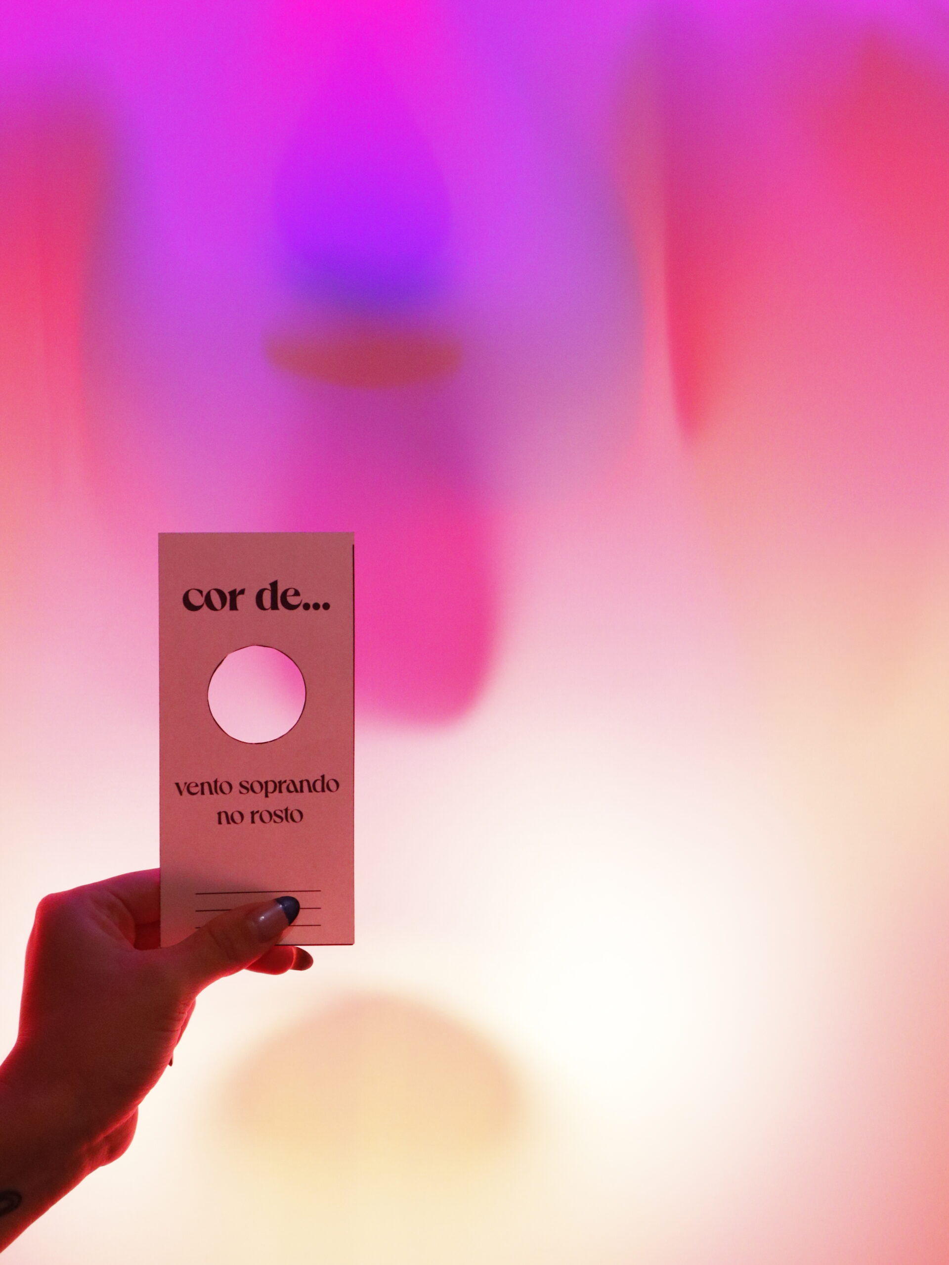 Descrição da imagem: Fotografia vertical de mão feminina segurando um pequeno cartão retangular. A mão é branca e a unha do polegar está pintada de azul-escuro na ponta superior, com o restante da unha em um tom cintilante claro. Segura o cartão pelo centro inferior. O cartão é branco e tem um recorte circular próximo ao topo. Acima do recorte, em letras pretas e negrito: “cor de...”. Abaixo do recorte, está o texto “vento soprando no rosto”. O fundo é desfocado, em tons de rosa, roxo, laranja e branco. Fim da descrição da imagem.
