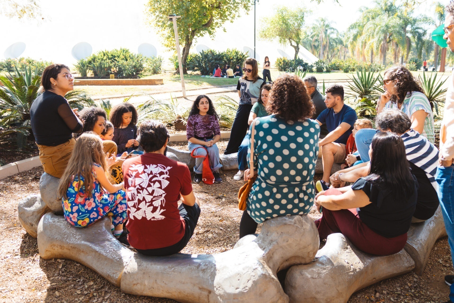 Descrição da imagem: Fotografia diurna horizontal da educadora Amanza Alves com um grupo de quinze pessoas ao ar livre no Parque Ibirapuera.
O grupo está em um pequeno espaço de terra batida, sentado em círculo, sobre estruturas de concreto cinza que se assemelham a enormes ossos estilizados.
Entre as pessoas, mulheres e homens adultos e crianças, com diferentes tons de pele; cabelos lisos, ondulados, cacheados e crespos; roupas em tons de azul, bege, marrom, vermelho, branco e preto. Amanza está sentada junto com o grupo e com o olhar voltado para as pessoas. Ela é negra de pele clara, de cabelos pretos cacheados, sobrancelhas médias, nariz e boca pequenos. Usa camiseta de mangas longas roxa, de tecido translúcido, e, por baixo, uma regata preta, calça jeans clara e tênis pretos.
Ao redor, há muitas árvores de copas esverdeadas e canteiros com plantas de folhas longas. Ao fundo, vista parcial da cúpula do edifício Oca. Fim da descrição da imagem.
