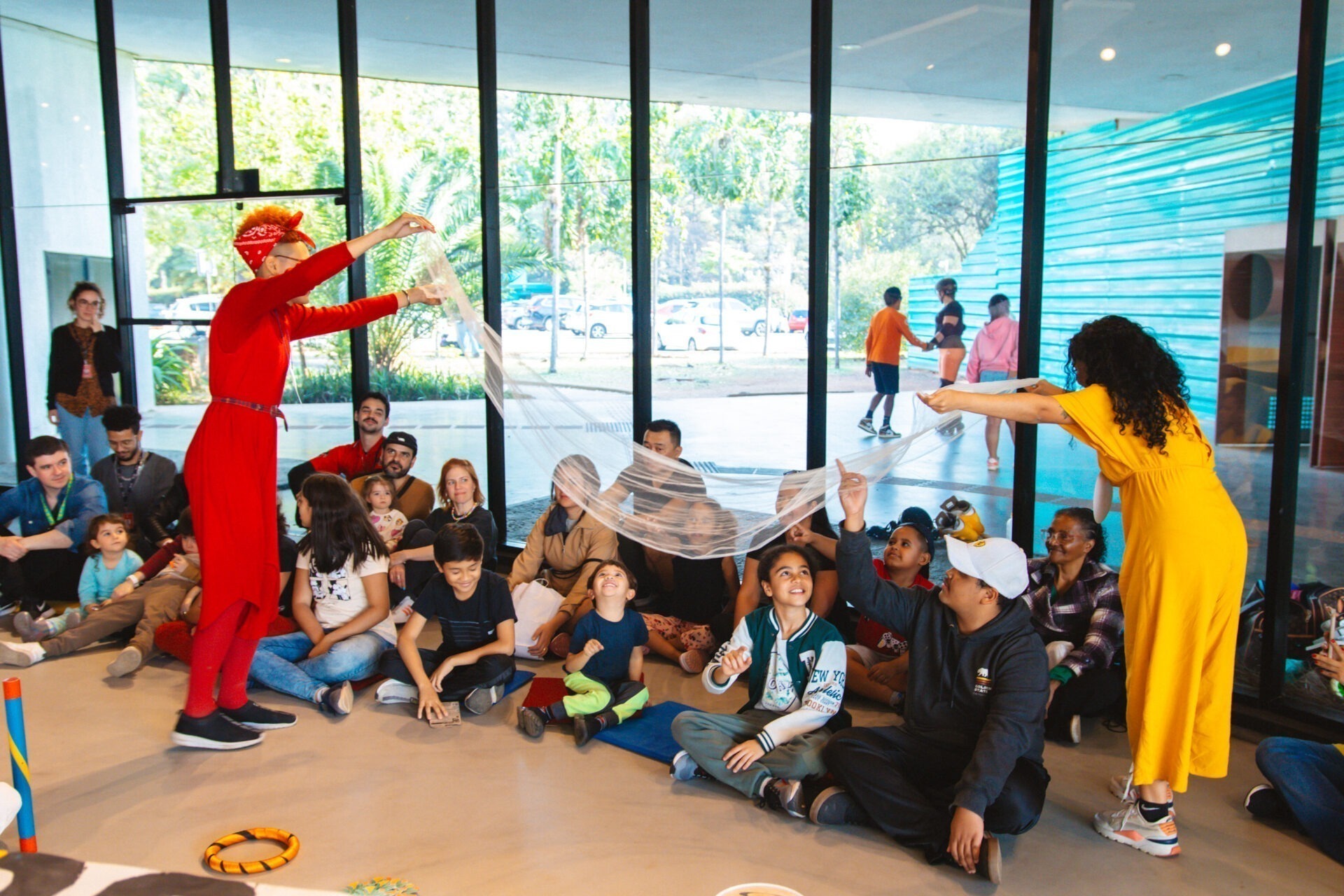 Descrição da imagem: Fotografia diurna horizontal das educadoras Luna e Amanda com um grupo de aproximadamente vinte pessoas sentadas no chão de um espaço interno amplo.
No grupo, há homens, mulheres e crianças. As educadoras estão em pé, seguram um tecido branco translúcido acima de algumas crianças do grupo, que sorriem. Outras crianças e adultos sentados observam o tecido elevado.
Luna está à esquerda. É uma mulher trans, negra de pele clara, tem cabelos ruivos, curtos, com as laterais raspadas. Usa um conjunto todo vermelho, uma bandana, uma segunda pele sob um macacão e tênis pretos. 
Amanda está à direita. É uma mulher negra de pele clara, de cabelos pretos cacheados. Usa macacão amarelo e tênis bege com pequenos detalhes cor de laranja.
Ao fundo, a parede é formada por painéis de vidro entremeados por esquadrias pretas, que deixam à mostra uma área externa com três pessoas juntas e árvores de copas verdes. Fim da descrição da imagem.

