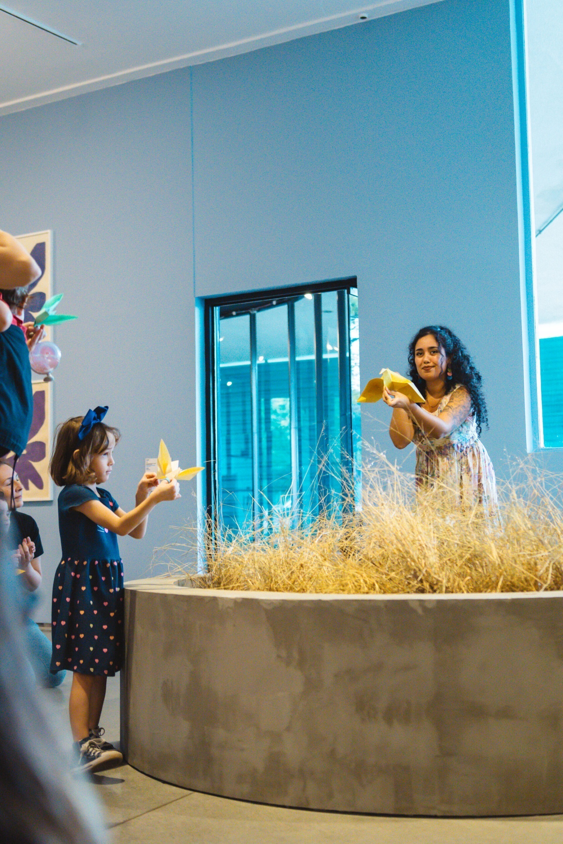 Descrição da imagem: Fotografia diurna vertical da educadora Amanza e uma menina em um ambiente interno, perto de uma instalação composta por uma mureta mediana de concreto em formato circular, preenchida com galhos secos. A menina está à esquerda, em pé, na lateral da estrutura. Ela tem aproximadamente sete anos, é branca, de cabelos lisos na altura dos ombros, sobrancelhas finas e nariz e boca pequenos. Usa laço azul na cabeça, vestido azul com a parte inferior estampada com corações cor-de-rosa, laranja e verde, e tênis escuros. Ela segura um origami amarelo em formato de pássaro Amanza está à direita, em pé, atrás da estrutura. Ela segura outro origami amarelo em formato de pássaro. Usa batom marrom e vestido branco florido em azul e roxo. Ao fundo, a parede é azul-claro. Fim da descrição da imagem.