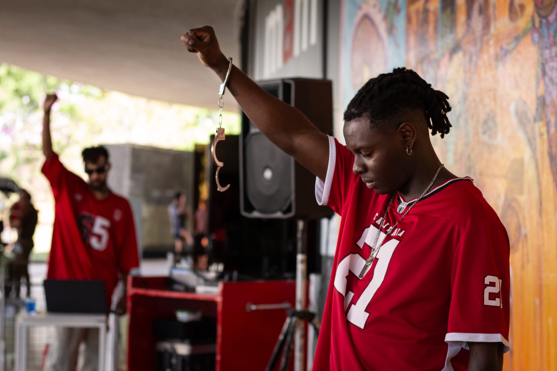 Descrição da imagem: Fotografia horizontal do artista Negret. Visto de perfil esquerdo, ele está em pé, à frente de uma parede com grafites cor de laranja. Está com a cabeça inclinada para baixo e os olhos fechados. O braço direito está erguido e a mão cerrada; no punho, há uma algema prateada, e a outra parte da algema está aberta e suspensa.
Ele é um homem negro retinto, de cabelos pretos com dreadlocks trançados para trás e laterais raspadas. As sobrancelhas são ralas, o nariz é arredondado na ponta e os lábios são volumosos. Usa camiseta vermelha com o número vinte e um estampado em branco na frente e também na lateral da manga. Usa brinco de argola pequeno e prateado e colar também prateado e curto. Ao fundo, em desfoque, há uma caixa de som e um Dj de óculos escuros com o olhar voltado para um notebook sobre uma pequena mesa branca. Fim da descrição da imagem.
