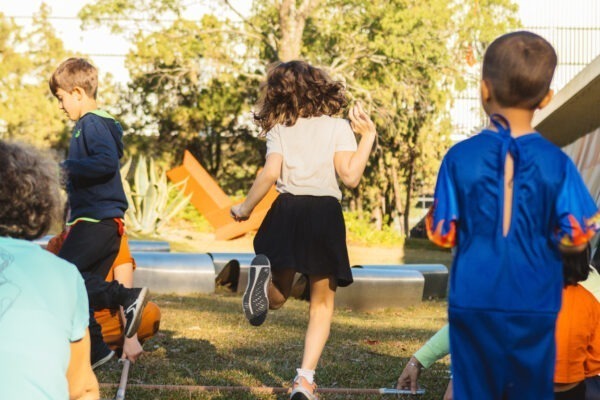 Imagem. Início da descrição de imagem: Fotografia capturada ao ar livre no Jardim de Esculturas, mostrando crianças em movimento durante uma atividade lúdica. Uma menina de camiseta branca e saia-shorts preto é vista de costas enquanto pula sobre varas de bambu dispostas no gramado. À direita, um menino usa uma fantasia de super-herói azul com detalhes de chamas. Ao fundo, árvores e a arquitetura do museu completam o cenário ensolarado. Fim da descrição de imagem.