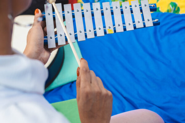 Imagem. Início da descrição de imagem: Fotografia colorida em plano médio e ângulo elevado, capturada por trás da figura de uma educadora do museu. Ela veste uma blusa branca texturizada e está sentada no chão, em frente a uma pessoa vestindo calça preta. A educadora segura um pequeno metalofone (xilofone com teclas de metal) azul com baquetas de madeira, tocando-o delicadamente. Ao fundo, observam-se tecidos coloridos estendidos no chão: um pano amarelo com estampas florais azuis e um pano azul sólido, criando um ambiente imersivo e lúdico. A imagem transmite uma sensação de tranquilidade, escuta atenta e exploração sensorial através do som. Fim da descrição de imagem.