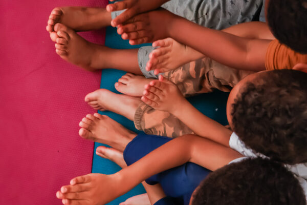 Imagem. Início da descrição de imagem: Fotografia colorida em plano médio e ângulo ligeiramente elevado, mostrando um grupo de crianças sentadas de forma descontraída sobre um tatame colorido (rosa e azul) em um ambiente de convivência do museu. A imagem foca no gesto das mãos das crianças, que estão estendidas à frente com as palmas unidas, parecendo realizar um gesto de saudação ou atenção durante uma atividade em grupo. Observam-se pés descalços e pernas cruzadas de diversas crianças, que vestem uniformes escolares claros e calças azuis. A iluminação é suave, transmitindo uma atmosfera de acolhimento e escuta atenta. Fim da descrição de imagem.