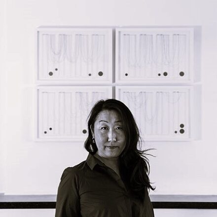 Imagem. Início da descrição de imagem: Retrato frontal em tom sépia de uma mulher de pele clara e ascendência asiática, com longos cabelos escuros e ondulados caídos sobre o ombro esquerdo. Ela olha diretamente para a câmera com uma expressão serena e um leve sorriso. Veste uma camisa escura de gola. A artista está posicionada centralizada à frente de um conjunto de quatro obras de arte emolduradas em branco, dispostas em uma grade de 2x2 na parede. As obras apresentam linhas finas curvas e pontos pretos de diferentes tamanhos sobre um fundo claro, evocando uma linguagem gráfica e minimalista. Abaixo das obras, vê-se uma linha horizontal escura que parece ser a borda de um móvel ou balcão. Fim da descrição de imagem.