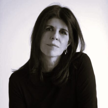 Imagem. Início da descrição de imagem: Retrato frontal em plano médio e tom sépia de uma mulher de pele clara, com longos cabelos escuros e lisos que emolduram seu rosto. Ela olha diretamente para a câmera com uma expressão serena, profunda e levemente melancólica. Usa brincos pequenos de metal com um design orgânico. Veste uma blusa escura de gola redonda e mangas compridas. O fundo da imagem é inteiramente branco e liso, criando um contraste suave que destaca a figura da artista, mantendo o foco em sua fisionomia e olhar introspectivo. Fim da descrição de imagem.