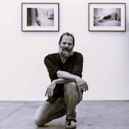Imagem. Início da descrição de imagem: Retrato frontal em tom sépia de um homem de pele clara, com cabelos escuros e uma barba longa e grisalha. Ele está agachado em uma perna só, com os braços cruzados sobre o joelho elevado, olhando diretamente para a câmera com uma expressão serena e profunda. Veste uma camisa escura de mangas compridas, calças jeans e sandálias. O artista está posicionado centralizado à frente de uma parede branca onde estão penduradas duas fotografias emolduradas: a da esquerda mostra uma queda d