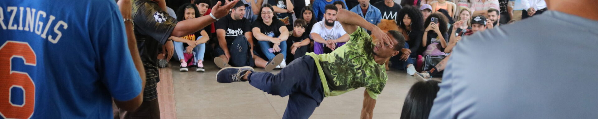 Imagem.
Início da descrição de imagem:
Fotografia colorida em plano aberto de uma roda de Breaking. No centro da roda, um jovem negro (B-boy) realiza um movimento de equilíbrio dinâmico: ele sustenta o corpo com apenas uma das mãos no chão, enquanto mantém as pernas elevadas e flexionadas no ar. Veste uma camiseta verde estampada, calça azul marinho e tênis esportivos azuis. Ao seu redor, uma grande plateia diversificada de jovens e adultos observa atentamente, muitos sentados no chão e outros em pé. Alguns espectadores registram a performance com seus celulares. A cena ocorre sob a icônica marquise de concreto branco projetada por Oscar Niemeyer, com grandes colunas em "V" visíveis ao fundo. A iluminação é natural e abundante, vinda das áreas abertas do parque.
Fim da descrição de imagem.