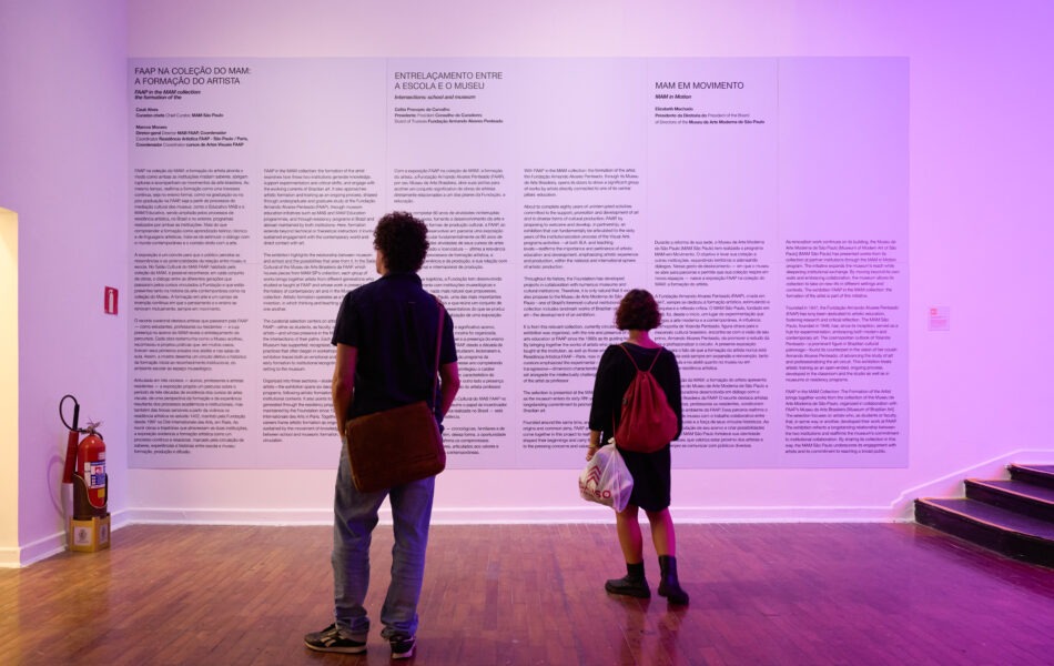Imagem. Descrição da imagem: Duas pessoas estão de costas em um espaço expositivo observando um grande painel branco com letras pretas fixado em uma parede iluminada com luz de coloração lilás. O painel contém textos explicativos da exposição "FAAP NA COLEÇÃO DO MAM: A FORMAÇÃO DO ARTISTA", organizados em colunas. À esquerda, um homem de cabelos cacheados, vestindo camisa azul marinho e calça jeans com uma bolsa tiracolo marrom. À direita, uma mulher com mochila vermelha e segurando uma sacola branca. No canto esquerdo, um extintor de incêndio está posicionado no chão próximo à parede. Fim da descrição da imagem.