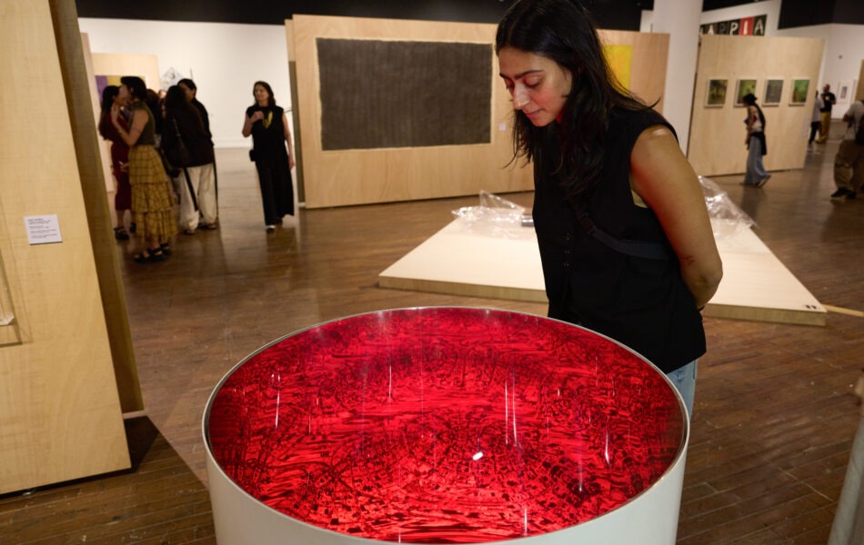 Imagem. Descrição da imagem: Uma mulher jovem de cabelos escuros e vestindo uma camiseta preta sem manga está inclinada, observando atentamente uma obra de arte circular. A obra é um recipiente cilíndrico branco com o topo preenchido por um líquido ou superfície reflexiva de cor vermelho intenso e brilhante, que apresenta padrões complexos de luz e sombra em seu interior. O ambiente da galeria tem piso de madeira e divisórias de compensado claro ao fundo, onde outras obras e visitantes circulam sob iluminação pontual de trilhos no teto. Fim da descrição da imagem.
