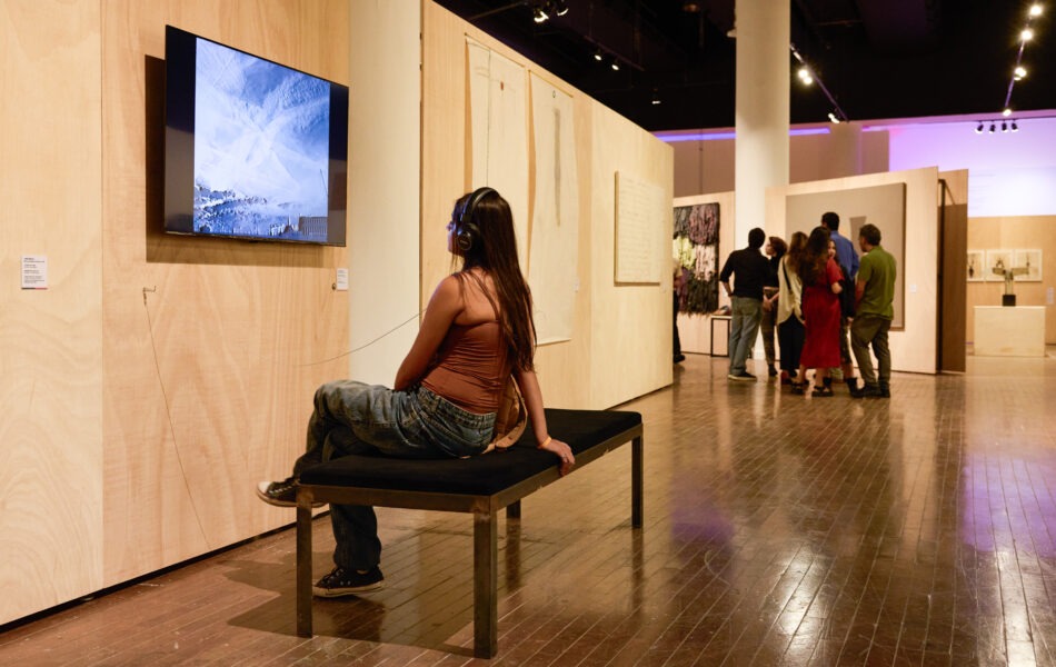 Imagem. Descrição da imagem: Uma jovem de cabelos longos, lisos e castanhos está sentada em um banco preto de estrutura metálica, usando fones de ouvido enquanto assiste a um vídeo em um monitor de TV fixado em um painel de madeira clara. O vídeo exibe uma imagem abstrata em tons de azul e branco. Ela veste uma blusa marrom de alças finas e calça jeans. Ao fundo, o espaço amplo da galeria revela outros painéis de madeira com pinturas e um grupo de pessoas conversando. A iluminação é quente e destaca o piso de madeira polida. Fim da descrição da imagem.