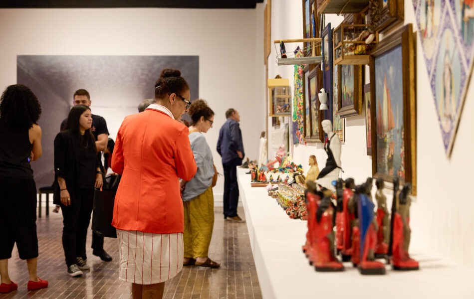 Imagem. Descrição da imagem: Vista lateral de uma exposição em uma galeria com piso de madeira. Em primeiro plano, uma mulher de pele retinta e cabelos presos em um coque alto, vestindo um blazer laranja vibrante sobre um vestido branco com listras vermelhas finas, caminha observando os itens expostos. À direita, sobre uma longa bancada branca que se estende para o fundo, há uma grande quantidade de pequenas esculturas coloridas e objetos. Na parede acima da bancada, vários quadros e caixas de madeira com miniaturas estão pendurados. Outros visitantes são vistos ao fundo, em um ambiente amplo e iluminado. Fim da descrição da imagem.