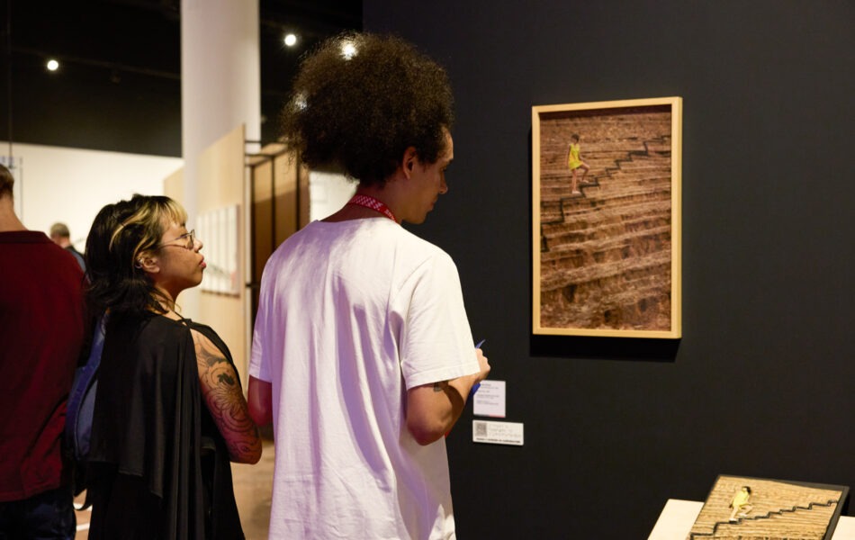 Imagem. Descrição da imagem: Em uma galeria de iluminação baixa, duas pessoas observam uma obra de arte fixada em uma parede preta. Em primeiro plano, um jovem de costas, com um cabelo afro volumoso e usando uma camiseta branca simples, foca sua atenção em uma fotografia vertical emoldurada em madeira clara. A fotografia retrata uma pessoa subindo uma escadaria de terra em uma encosta. Ao lado do jovem, à esquerda, uma mulher de óculos e cabelos escuros com mechas claras observa a mesma obra. No canto inferior direito, há uma pequena maquete ou relevo no chão que parece reproduzir a escadaria vista na foto. Fim da descrição da imagem.