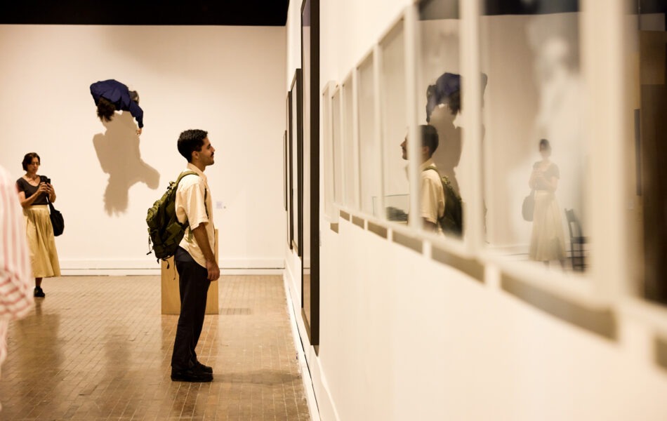 Imagem. Descrição da imagem: Em uma galeria de arte com piso de madeira e paredes brancas, um homem de pele clara e cabelos escuros, usando uma camisa bege e uma mochila verde militar grande, está em pé de perfil observando obras na parede à sua frente. Ele olha para uma sequência de quadros com molduras brancas e vidros reflexivos que ocupam o lado direito do quadro, onde se vê o seu próprio reflexo e o de uma mulher ao fundo. Ao fundo, no centro da galeria, uma escultura azul em formato de corpo humano parece flutuar ou estar fixada no alto da parede branca, projetando uma sombra marcante. À esquerda, outra mulher está de pé fotografando a cena com um celular. Fim da descrição da imagem.