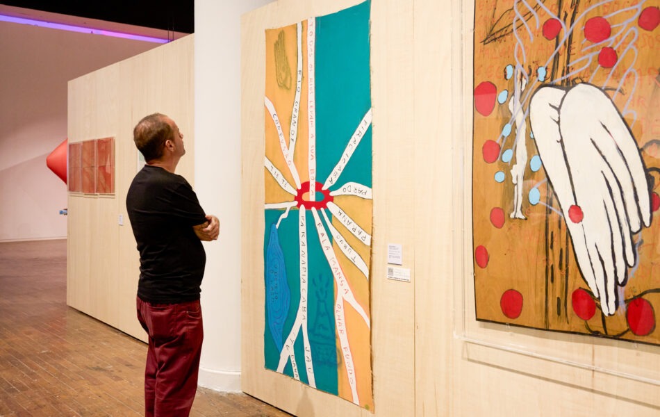 Imagem. Descrição da imagem: Um homem de costas, vestindo camiseta preta e calça vinho, está com os braços cruzados observando obras de arte montadas em grandes painéis de madeira compensada clara. À sua frente, um painel vertical exibe uma pintura colorida com formas que lembram pétalas ou raios saindo de um centro vermelho, contendo nomes escritos. À direita, outro painel mostra uma obra com o desenho de um par de asas brancas sobre um fundo ocre pontuado por círculos vermelhos. O piso é de madeira escura e o teto da galeria é preto, com luzes embutidas. Fim da descrição da imagem.