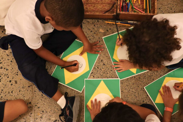 Imagem. Início da descrição de imagem: Fotografia colorida em plano zenital (visto de cima) de cinco crianças sentadas em roda no chão de granilite da marquise do Ibirapuera. Elas estão debruçadas sobre folhas de papel que reproduzem o padrão geométrico da bandeira do Brasil (losango amarelo sobre retângulo verde). Cada criança utiliza um lápis grafite para desenhar um rosto ou autorretrato dentro do círculo central branco da bandeira, onde originalmente estaria o céu estrelado. Ao fundo, observam-se caixas de madeira repletas de lápis de cor coloridos, sugerindo o contexto de uma oficina pedagógica. Fim da descrição de imagem.