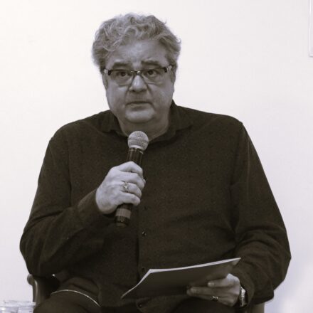 Início da descrição de imagem: Retrato frontal em tom sépia de um homem de pele clara, com cabelos grisalhos e cacheados. Ele usa óculos de grau com armação escura e olha para frente com uma expressão atenta. O homem está sentado, vestindo uma camisa de botões escura, e segura um microfone com a mão direita, enquanto a mão esquerda apoia papéis em seu colo. Em seus dedos, nota-se o uso de anéis, e no pulso esquerdo, um relógio. O fundo da imagem é uma parede clara e lisa, característica de um ambiente de debate ou palestra. Fim da descrição de imagem.