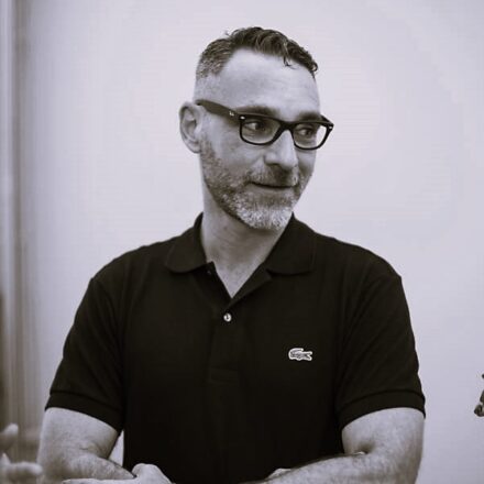 Imagem. Retrato em tom sépia de um homem de pele clara, com cabelos escuros curtos e barba grisalha bem aparada. Ele olha para o lado direito da câmera com uma expressão atenta e um leve sorriso. Usa óculos de grau com armação escura e marcante. Veste uma camisa polo escura com um pequeno logotipo bordado no peito. O artista está com os braços cruzados. O fundo da imagem é claro e liso, mantendo o foco total na figura do artista e em sua expressão comunicativa. Fim da descrição de imagem.