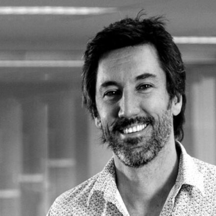 Imagem. Início da descrição de imagem: Retrato frontal em preto e branco de um homem de pele clara, com cabelos escuros e lisos, levemente arrepiados, barba e bigode bem aparados. Ele olha diretamente para a câmera com um sorriso aberto e expressivo. Veste uma camisa clara com uma padronagem de pequenas estampas repetitivas. O fundo da imagem está desfocado, sugerindo um ambiente interno iluminado. Fim da descrição de imagem.