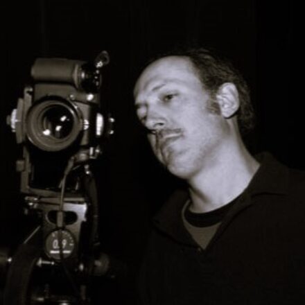 Imagem. Início da descrição de imagem: Retrato em tom sépia, capturado em ângulo médio, de um homem de pele clara com cabelos curtos e escuros nas laterais e calvície frontal. Ele possui um bigode fino e olha atentamente para o visor de uma câmera fotográfica profissional de grande formato, montada em um tripé à sua esquerda. O homem veste uma camisa polo escura sobre uma camiseta de gola redonda. O fundo da imagem é inteiramente preto e liso, destacando a figura do fotógrafo e seu equipamento em um ambiente que sugere um estúdio. Fim da descrição de imagem.