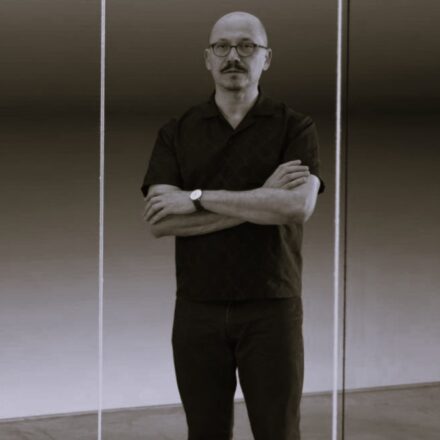 Imagem. Início da descrição de imagem: Retrato frontal em tom sépia de um homem calvo, de pele clara, com bigode e cavanhaque escuros. Ele olha diretamente para a câmera com uma expressão séria e braços cruzados na altura do peito. Usa óculos de grau com armação escura e arredondada, e um relógio de pulso com pulseira escura no braço esquerdo. Veste uma camisa de botões escura com uma textura sutil de padronagem geométrica e calças escuras. O artista está posicionado entre duas linhas verticais claras e finas, que parecem ser reflexos ou elementos de uma instalação em vidro, criando uma composição simétrica e rigorosa. O fundo é neutro e suavemente iluminado. Fim da descrição de imagem.