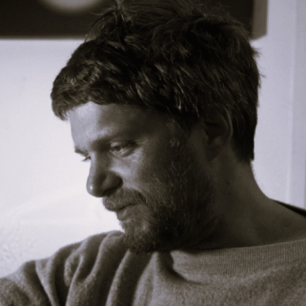 Imagem. Início da descrição de imagem: Retrato em close-up e tom sépia de um homem de pele clara, com cabelos escuros curtos e desalinhados, e uma barba e bigode castanhos cheios. Ele está posicionado de perfil, olhando para baixo e para a esquerda, com uma expressão concentrada e introspectiva. Veste um blusão de lã ou moletom de textura grossa e gola redonda. A iluminação é dramática, vinda da esquerda, o que cria um forte contraste de luz e sombra (chiaroscuro) em seu rosto, deixando parte de sua fisionomia mergulhada na escuridão. O fundo é neutro e desfocado, mantendo o foco absoluto no semblante do artista. Fim da descrição de imagem.