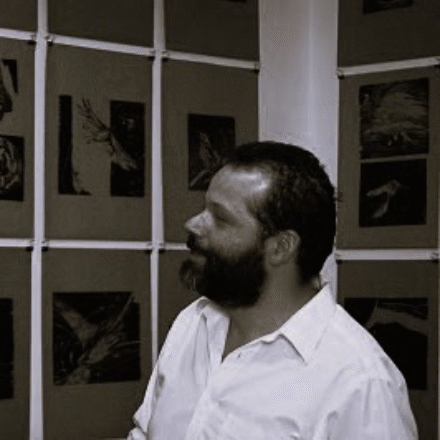 Imagem. Início da descrição de imagem: Retrato em tom sépia, em plano médio e perfil esquerdo, de um homem de pele clara, com cabelos escuros e uma barba e bigode cheios e bem desenhados. Ele olha para a frente com uma expressão contemplativa. Veste uma camisa social clara de mangas compridas, levemente desabotoada no colarinho. O artista está posicionado à frente de uma parede que exibe suas obras: uma série de xilogravuras de pequenos formatos, fixadas em painéis escuros separados por ripas claras verticais e horizontais. As gravuras apresentam formas orgânicas e silhuetas de pássaros e plantas. A iluminação lateral destaca os contornos de seu rosto e a textura das obras ao fundo. Fim da descrição de imagem.