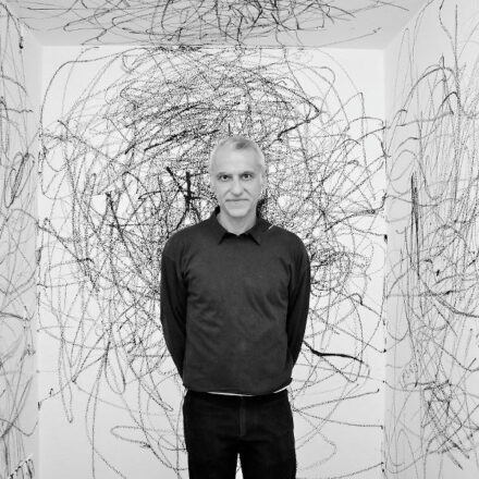 Imagem. Início da descrição de imagem: Retrato frontal em preto e branco de um homem de pele clara, com cabelos curtos e grisalhos. Ele olha diretamente para a câmera com uma expressão serena e um leve sorriso. O artista está de pé, com os braços atrás do corpo, vestindo uma blusa de manga longa escura sobre uma camiseta clara e calças pretas. Ele está posicionado no centro de um ambiente de paredes brancas totalmente cobertas por desenhos abstratos de linhas pretas frenéticas e emaranhadas, que lembram rabiscos em larga escala feitos com carvão ou grafite. Fim da descrição de imagem.