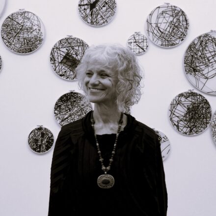 Imagem. Início da descrição de imagem: Retrato frontal em preto e branco de uma mulher de pele clara, com cabelos brancos, curtos e cacheados. Ela olha para o lado direito da câmera com um sorriso sereno. Veste uma blusa escura e um colar longo com um pingente oval grande e detalhado. A artista está posicionada à frente de uma parede branca onde estão instaladas diversas obras circulares de diferentes tamanhos, que contêm desenhos complexos de linhas pretas emaranhadas, lembrando tramas ou mapas abstratos. Fim da descrição de imagem.