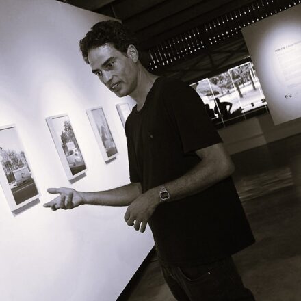Imagem. Início da descrição de imagem: Retrato em tom sépia, capturado em um ângulo dinâmico e levemente inclinado (plano holandês), de um homem jovem de pele clara com cabelos escuros e cacheados. Ele está posicionado em plano médio, vestindo uma camiseta escura de gola redonda e calças jeans, com um relógio de pulso metálico e claro visível no braço esquerdo. O artista olha para a esquerda e estende a mão direita em um gesto explicativo, como se estivesse em meio a uma conversa ou apresentação. Ele está em pé em um ambiente de galeria; ao fundo, fixadas em uma parede branca, veem-se três fotografias emolduradas em branco dispostas em uma linha horizontal, que remetem a documentações de arquiteturas urbanas. Mais ao fundo, através de uma abertura envidraçada, observa-se a paisagem do Parque Ibirapuera, o que localiza a cena no MAM São Paulo. O chão é de cimento polido e o ambiente é banhado por luz natural e artificial. Fim da descrição de imagem.
