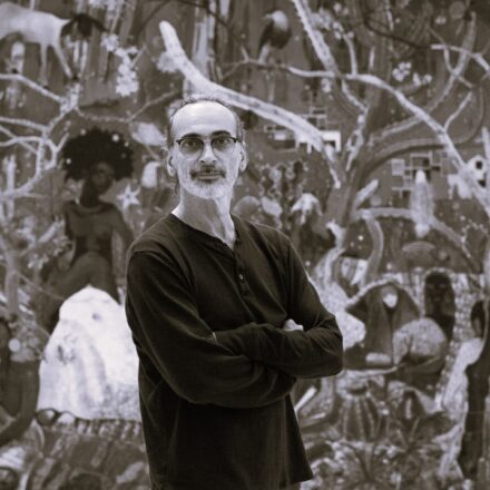 Imagem. Início da descrição de imagem: Retrato frontal em tom sépia de um homem de pele clara, com cabelos escuros presos para trás e uma barba grisalha curta e bem aparada. Ele olha diretamente para a câmera com uma expressão serena e braços cruzados na altura do peito. Usa óculos de grau com armação escura e arredondada e veste uma camiseta de gola "henley" escura de mangas compridas. O artista está posicionado em primeiro plano, à frente de uma de suas pinturas monumentais que ocupa todo o fundo. A obra ao fundo exibe uma densa composição de formas orgânicas, galhos de árvores, figuras humanas e animais, integrados em uma narrativa visual rica em detalhes e texturas, característica de sua pesquisa sobre a natureza e a cultura. Fim da descrição de imagem.