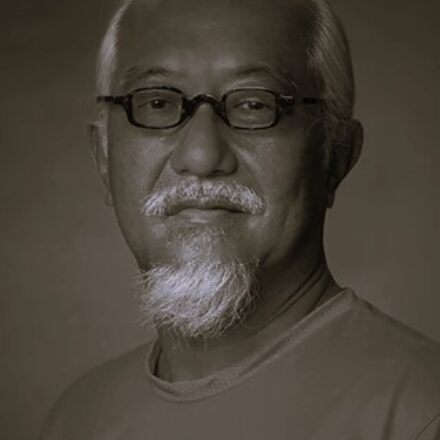 Imagem. Início da descrição de imagem: Retrato frontal em tom sépia, em plano fechado (close-up), de um homem de pele clara e ascendência asiática. Ele possui cabelos grisalhos curtos, bigode e um cavanhaque longo e cheio, também grisalhos. Olha diretamente para a câmera com uma expressão serena e contemplativa através de óculos de grau com armação escura e arredondada. Veste uma camiseta de gola redonda em tom médio. O fundo da imagem é neutro e desfocado, concentrando toda a atenção nas feições do artista. Fim da descrição de imagem.