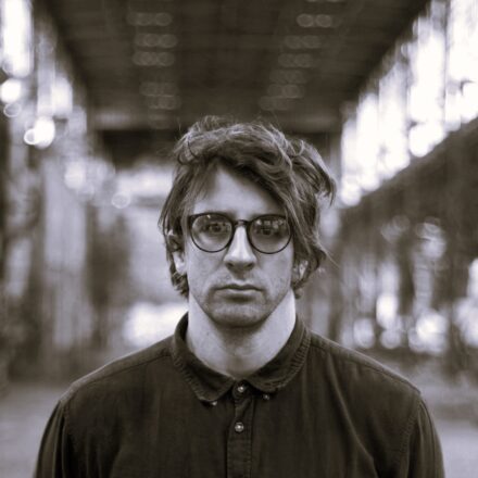 Imagem. Início da descrição de imagem: Retrato frontal em tom sépia de um homem de pele clara, com cabelos escuros e levemente bagunçados. Ele olha diretamente para a câmera através de óculos de grau com armação redonda e escura, apresentando uma expressão séria e neutra. Veste uma camisa de botões escura, fechada até o colarinho. O fundo da imagem está desfocado, mas revela uma estrutura arquitetônica industrial com teto alto e vigas metálicas, sugerindo um galpão ou um grande pavilhão. Fim da descrição de imagem.