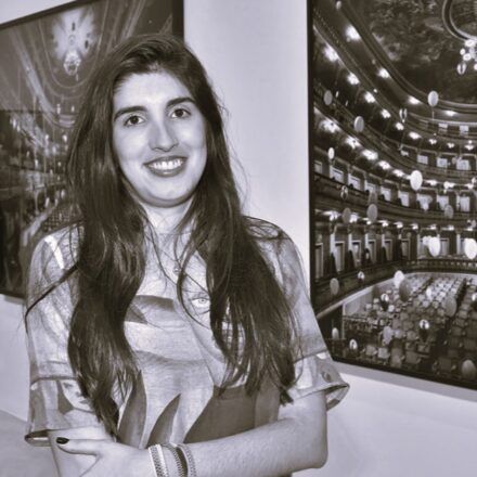 Imagem. Início da descrição de imagem: Retrato frontal em tom sépia de uma mulher de pele clara, com longos cabelos escuros e ondulados que caem sobre seus ombros. Ela olha diretamente para a câmera com um sorriso aberto. A artista está com os braços cruzados, vestindo uma blusa estampada, e usa pulseiras no pulso direito. Ela está posicionada entre duas de suas obras fotográficas emolduradas, que retratam interiores de teatros históricos suntuosos repletos de balões flutuantes, uma marca característica de sua produção artística. Fim da descrição de imagem.