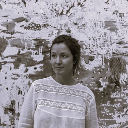 Imagem. Início da descrição de imagem: Retrato frontal em plano médio e tom sépia de uma mulher de pele clara, com cabelos escuros presos para trás. Ela olha para o lado esquerdo da câmera com uma expressão serena e observadora. Veste uma blusa clara com detalhes de renda horizontal (entremeios) no busto e nas mangas, conferindo uma textura delicada ao traje. A artista está posicionada em primeiro plano, à frente de uma de suas pinturas de grande escala que ocupa todo o fundo. A obra ao fundo apresenta uma composição abstrata e matérica, com pinceladas expressivas e sobreposições de camadas que sugerem uma paisagem fragmentada, característica marcante de sua produção pictórica. Fim da descrição de imagem.