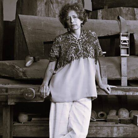 Imagem. Início da descrição de imagem: Retrato frontal em tom sépia da artista Elisa Bracher. Ela é uma mulher de cabelos escuros, curtos e cacheados, e olha diretamente para a câmera com uma expressão serena e compenetrada. Veste uma blusa solta com estampas étnicas, florais ou geométricas na parte superior e calças claras com padrão xadrez suave. A artista está apoiada em uma bancada de madeira robusta, e o fundo da imagem mostra grandes estruturas de madeira brutas e empilhadas, possivelmente parte de suas esculturas ou instalações em processo, além de objetos cerâmicos e ferramentas em prateleiras inferiores. Fim da descrição de imagem.