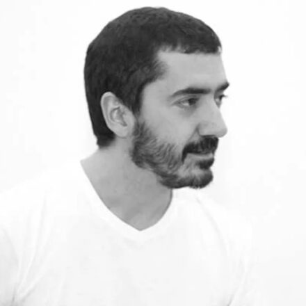 Imagem.Início da descrição de imagem: Retrato em preto e branco de um homem de pele clara, com cabelos curtos e escuros, barba e bigode bem definidos. Ele está posicionado de perfil parcial, olhando para a direita da câmera com uma expressão pensativa e serena. Veste uma camiseta branca simples de gola redonda. O fundo da imagem é inteiramente branco e liso, criando um contraste nítido que destaca os contornos do rosto e do cabelo do artista em uma estética minimalista. Fim da descrição de imagem.