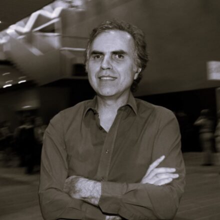 Imagem. Início da descrição de imagem: Retrato em tom sépia, em plano médio, de um homem de pele clara, com cabelos escuros e levemente grisalhos nas laterais. Ele olha diretamente para a câmera com uma expressão serena e um leve sorriso. Está com os braços cruzados na altura do peito, vestindo uma camisa de botões de cor escura e mangas compridas. O artista está posicionado em um ambiente interno amplo; ao fundo, à esquerda, observa-se uma escadaria e uma estrutura arquitetônica moderna e dinâmica. O fundo apresenta um leve desfoque de movimento, o que confere profundidade à cena e mantém o foco principal na figura do artista. Fim da descrição de imagem.