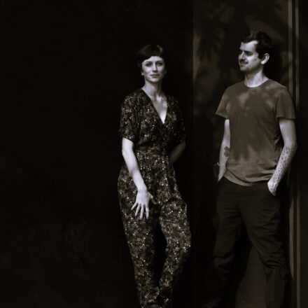 Imagem. Início da descrição de imagem: Retrato em tom sépia de um homem e uma mulher posicionados contra uma parede escura. À esquerda, a mulher está de frente, com uma das mãos no quadril, vestindo um macacão estampado de mangas curtas e decote em "V". Ela possui cabelos curtos e escuros. À direita, o homem está encostado na parede, olhando em direção à mulher. Ele veste uma camiseta de mangas curtas e calças escuras, exibindo tatuagens geométricas nos antebraços. Sombras de folhas de plantas são projetadas sobre a parede e sobre o homem, criando um jogo de luz e sombra na composição. Fim da descrição de imagem.