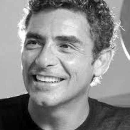 Imagem. Início da descrição de imagem: Retrato em close-up, em preto e branco, de um homem de pele clara com cabelos escuros, curtos e cacheados. Ele está posicionado levemente de perfil, olhando para cima e para a esquerda, com um sorriso aberto e radiante que mostra os dentes. Veste uma camiseta escura de gola redonda. A iluminação é suave, destacando suas feições e o brilho nos olhos, com um fundo neutro e claro que mantém o foco total em sua expressão entusiasmada. Fim da descrição de imagem.