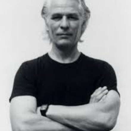 Imagem. Início da descrição de imagem: Retrato frontal em preto e branco de um homem de pele clara, com cabelos grisalhos penteados para trás. Ele olha diretamente para a câmera com uma expressão serena e braços cruzados na altura do peito, evidenciando um relógio de pulso escuro no braço esquerdo. Veste uma camiseta preta simples de gola redonda. O fundo da imagem é inteiramente branco e liso, criando um contraste marcante que destaca a figura do artista de forma minimalista e atemporal. Fim da descrição de imagem.