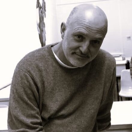 Imagem. Início da descrição de imagem: Retrato frontal em preto e branco de um homem calvo de pele clara, com barba e bigode grisalhos. Ele olha diretamente para a câmera com uma expressão serena e um leve sorriso. Veste um suéter de lã cinza texturizado sobre uma camiseta branca de gola redonda. O fundo da imagem está desfocado, revelando um ambiente interno claro com o que parece ser uma mesa de trabalho e cadeiras brancas ao fundo, sugerindo um ateliê ou escritório. À esquerda, vê-se a silhueta desfocada de uma obra de arte ou objeto. Fim da descrição de imagem.