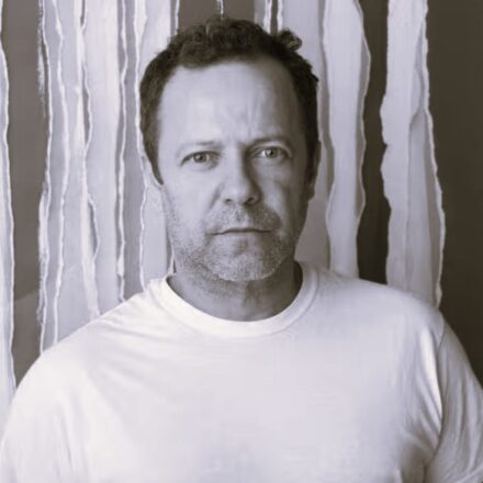 Imagem. Início da descrição de imagem: Retrato frontal em plano médio e tom sépia de um homem de pele clara, com cabelos escuros levemente ondulados e barba grisalha por fazer. Ele olha diretamente para a câmera com uma expressão séria e penetrante. Veste uma camiseta branca simples de gola redonda. O artista está posicionado centralizado à frente de um fundo composto por faixas verticais claras de bordas irregulares e rasgadas, que criam um padrão rítmico e sugerem uma de suas obras de intervenção ou estudo de materialidade. A iluminação é frontal e suave, destacando os detalhes de sua fisionomia e a textura do fundo. Fim da descrição de imagem.