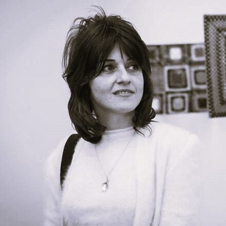 Imagem. Início da descrição de imagem: Retrato em tom sépia de uma mulher de pele clara, com cabelos lisos escuros, volumosos e com franja, na altura dos ombros. Ela olha para a direita da câmera com uma expressão suave e um leve sorriso. Veste uma blusa de lã clara e felpuda, usa um colar fino com um pingente em formato de gota e carrega uma bolsa com alça escura sobre o ombro direito. Ao fundo, à direita, veem-se fragmentos de obras de arte com padronagens quadriculadas e texturas que remetem ao universo têxtil, típicas de suas produções que exploram o suporte e a materialidade. Fim da descrição de imagem.