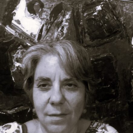 Imagem. Início da descrição de imagem: Retrato em tom sépia, em plano médio (close-up), de uma mulher de pele clara, com cabelos grisalhos curtos, lisos e levemente bagunçados. Ela olha diretamente para a câmera com uma expressão serena e contemplativa. Veste uma blusa estampada com uma gola de padronagem complexa e veste brincos claros de formato arredondado. A artista está posicionada em primeiro plano, à frente de uma obra de arte abstrata e texturizada que ocupa todo o fundo. A obra de fundo apresenta formas densas e pinceladas expressivas em tons de sépia, com um fragmento de forma orgânica clara no canto superior esquerdo, criando uma atmosfera que mimetiza o estilo de suas próprias criações. Fim da descrição de imagem.