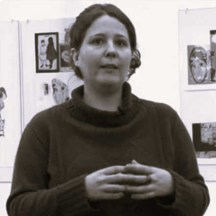Imagem. Início da descrição de imagem: Retrato frontal em plano médio, capturado em tom sépia, de uma mulher de pele clara. Ela possui cabelos escuros ondulados, presos para trás. Seu olhar está voltado levemente para cima e para a direita, com a boca semiaberta, sugerindo que está em meio a uma fala ou explicação. Veste um suéter de lã escuro com gola alta texturizada e padronagem de tranças. Suas mãos estão posicionadas à frente do corpo, com as pontas dos dedos tocando-se, um gesto comum de argumentação. Ao fundo, em uma parede clara, veem-se diversas obras de arte fotográficas e gráficas emolduradas, desfocadas, indicando um ambiente de exposição ou auditório. Fim da descrição de imagem.